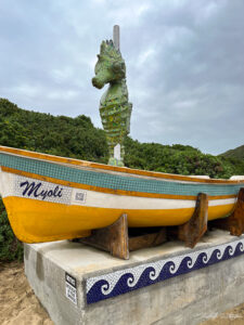 A mosaiced yellow boat named "Myoli" displayed on a pedestal with a mosaic Knysna seahorse statue in the background.