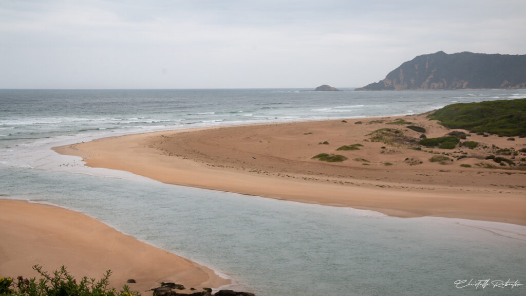 A wide view of a coastal landscape with a sandy beach curving along the shoreline, a river flowing into the ocean, and hills in the background