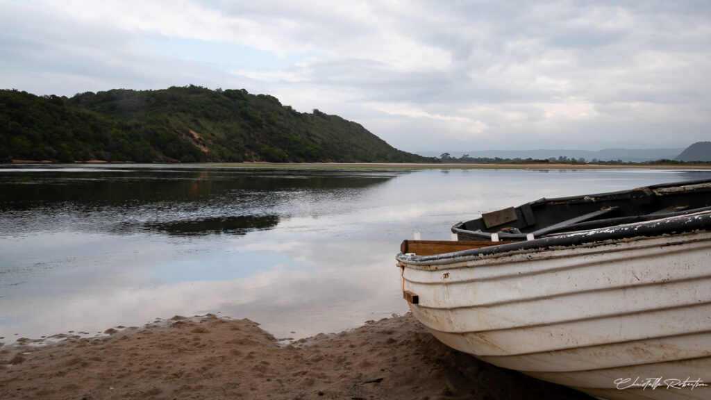 A calm river with green hills in the background and an old boat resting on the sandy shore.