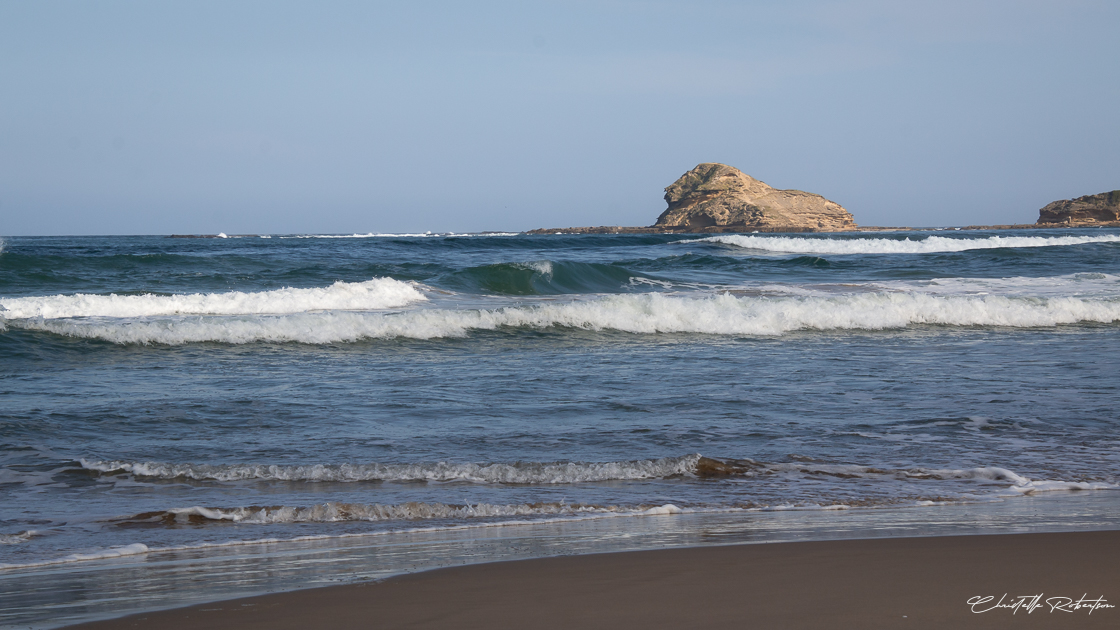 A coastal scene with waves crashing onto the shore and a Sphinx-like sandstone rock formation in the distance under a clear sky.