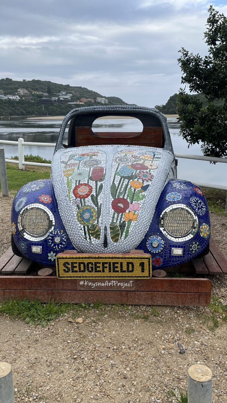 A decorated Volkswagen Beetle car front, covered in colorful mosaic tiles with floral designs, labeled "SEDGEFIELD 1."